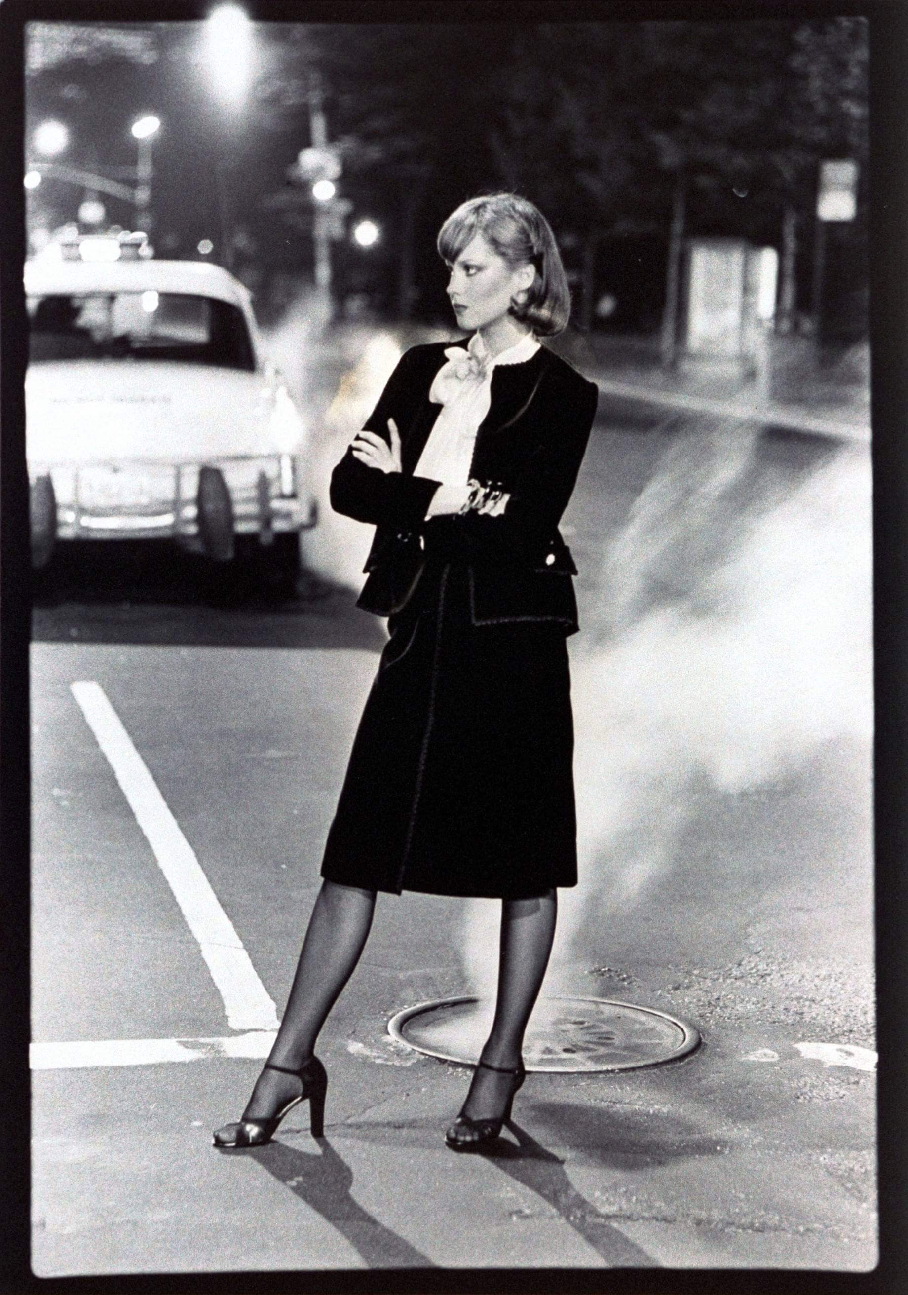 Model Rosie Vela standing with arms crossed in the street in New York City, near a steaming manhole Vogue cover, wearing a black velvet Chanel suit with wrap skirt and a small jacket over white satin blouse, by Belle Saunders for Abe Schrader, 1975. (Guliver Photos/ Getty Images).